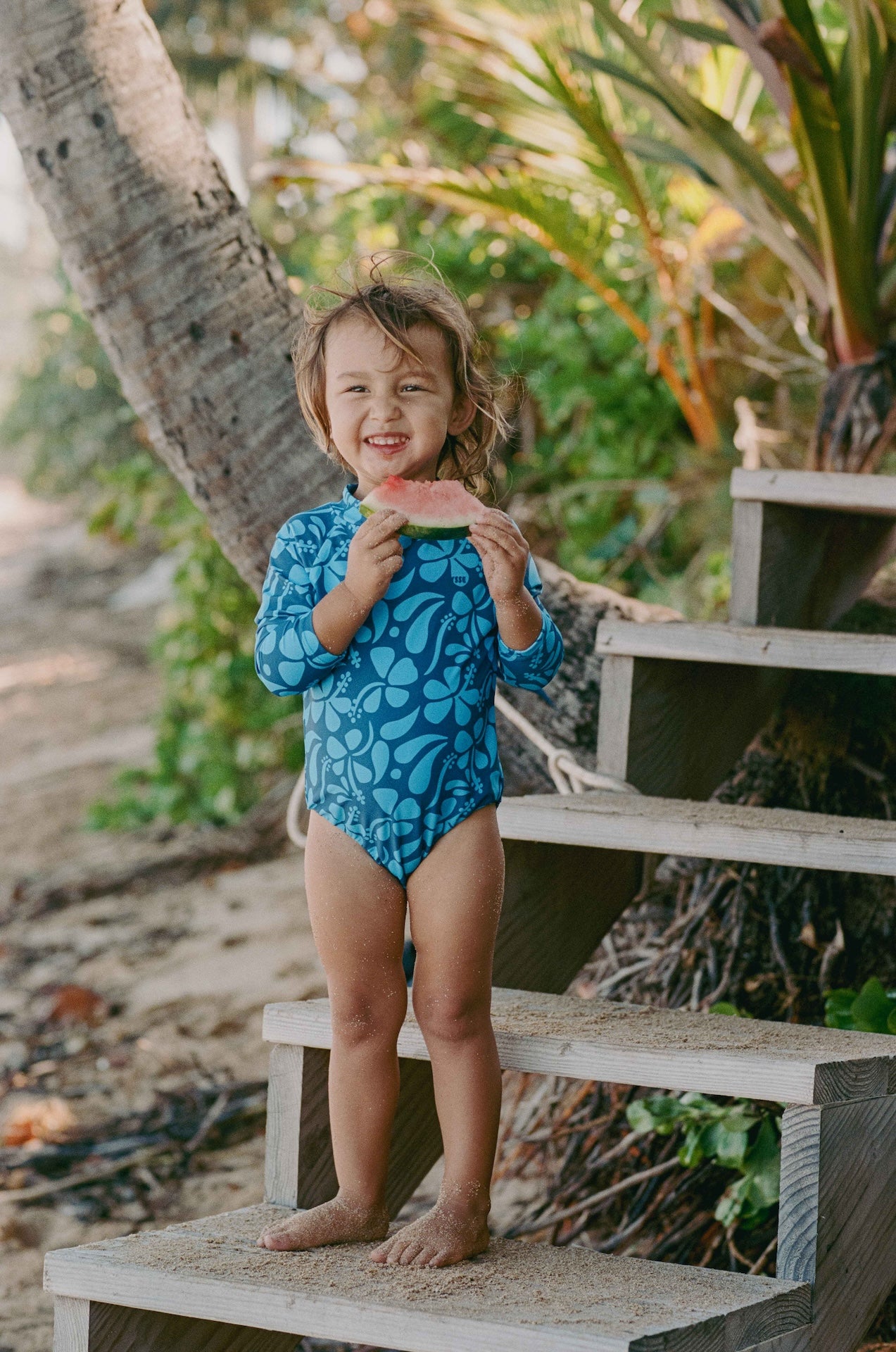 Child in a blue swimsuit holding watermelon on a wooden platform with greenery in the background