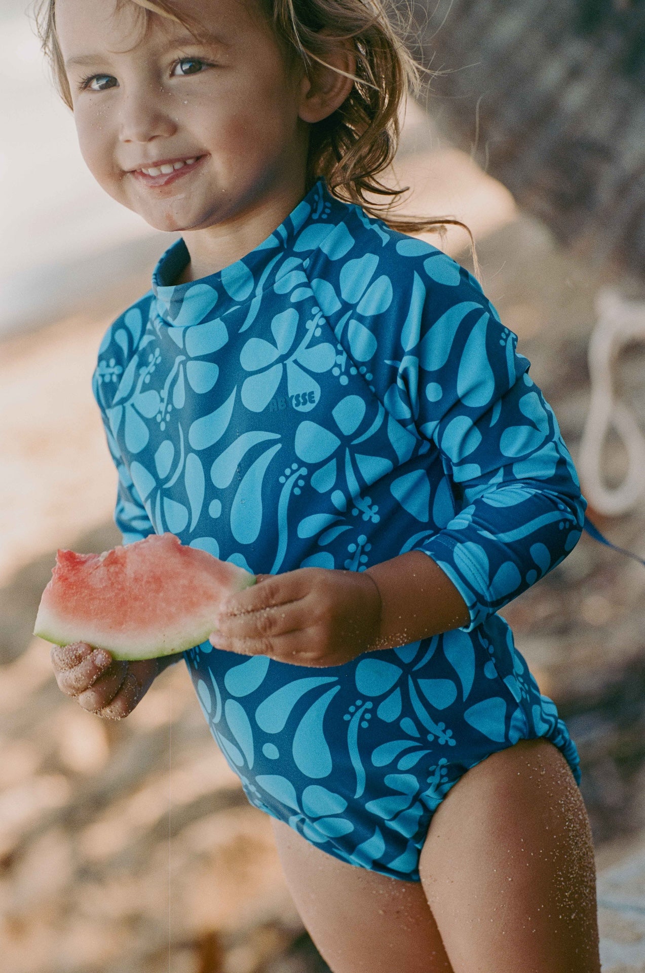 Child in a blue floral swimsuit eating watermelon on a beach.