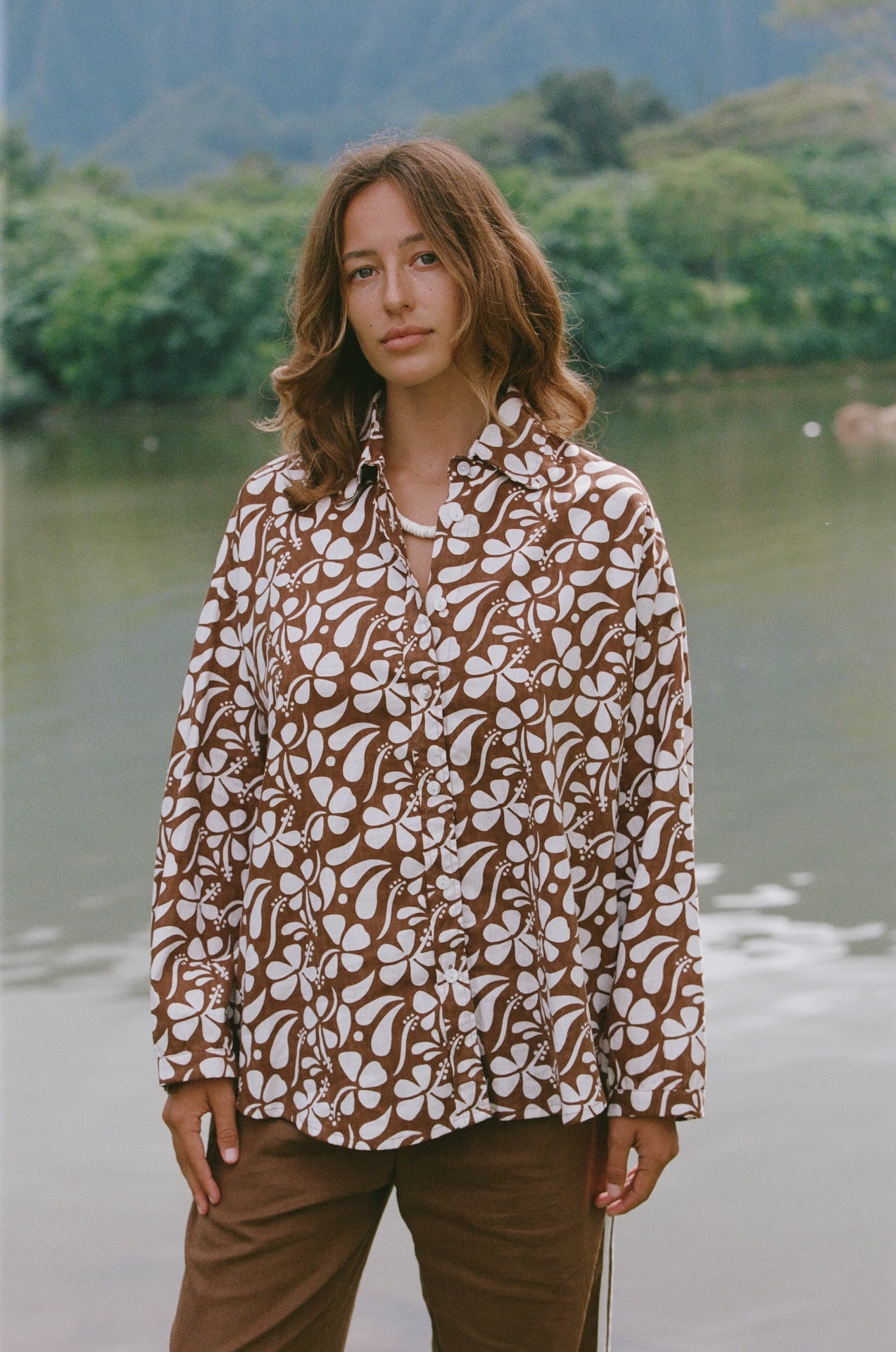Woman wearing a patterned shirt standing by a body of water with mountains in the background