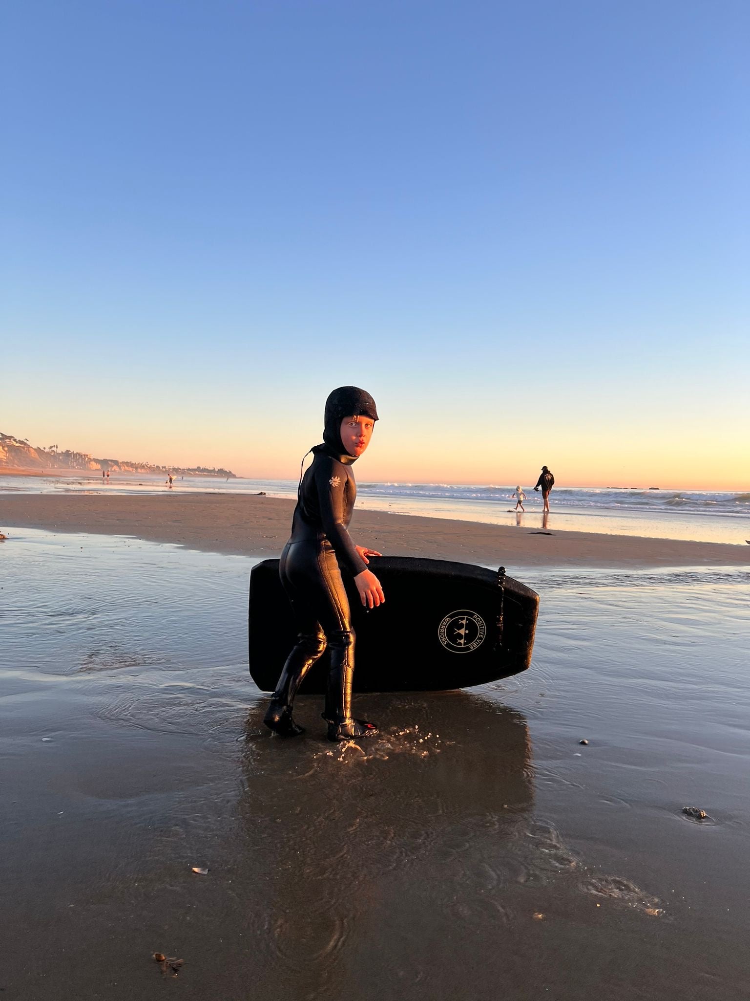 Child in a wetsuit sitting on a bodyboard at sunset on a beach