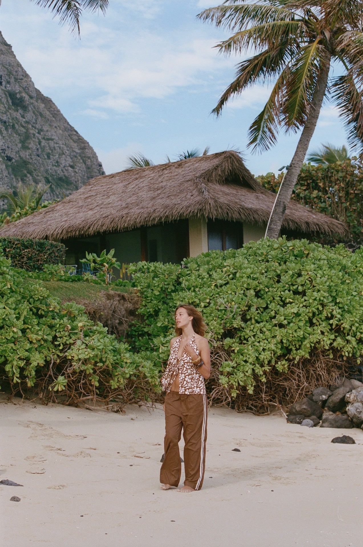 Person standing on a sandy beach with a thatched-roof hut and palm trees in the background.