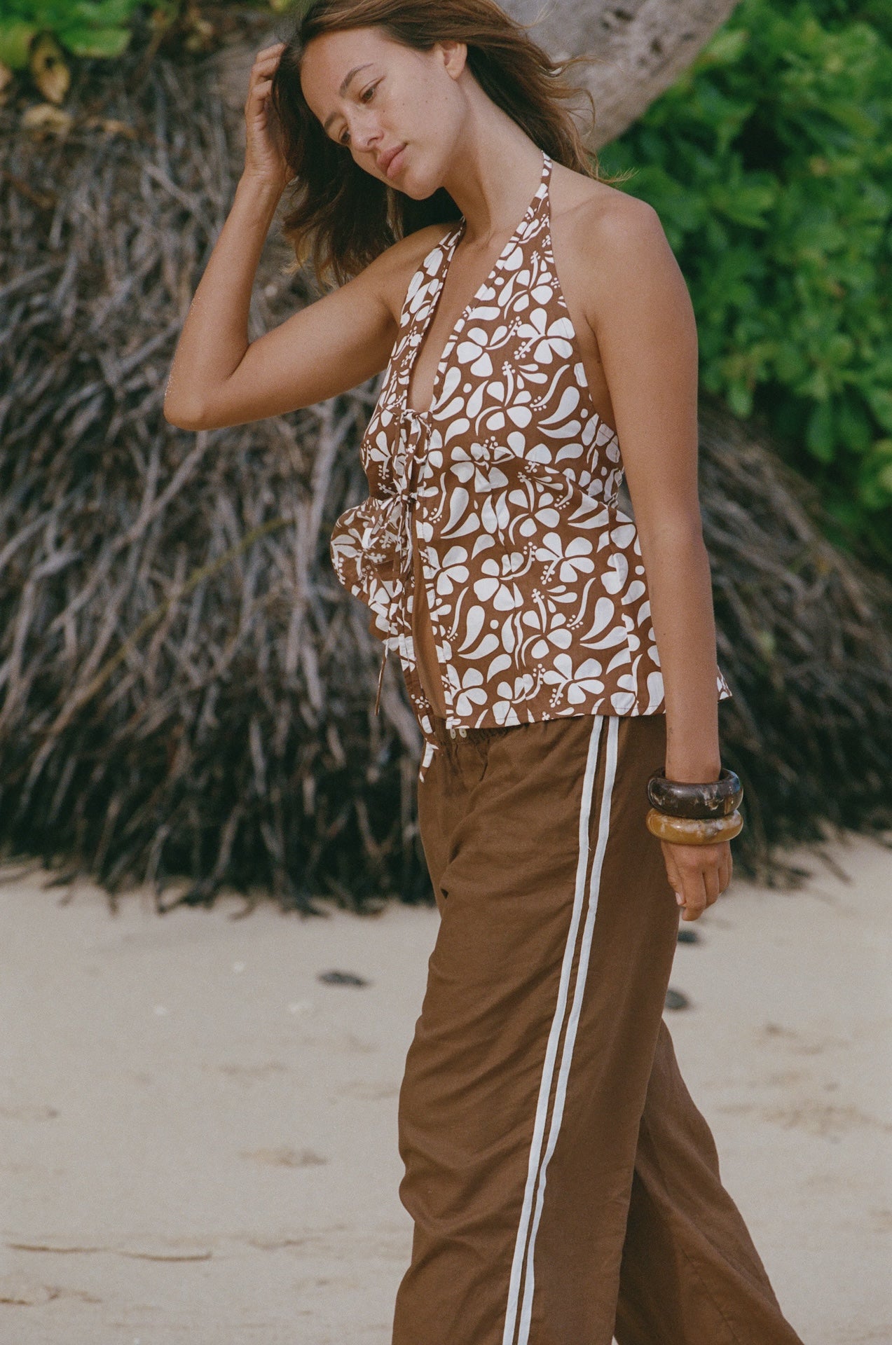 Woman in a patterned top and brown pants with white stripes on a beach.
