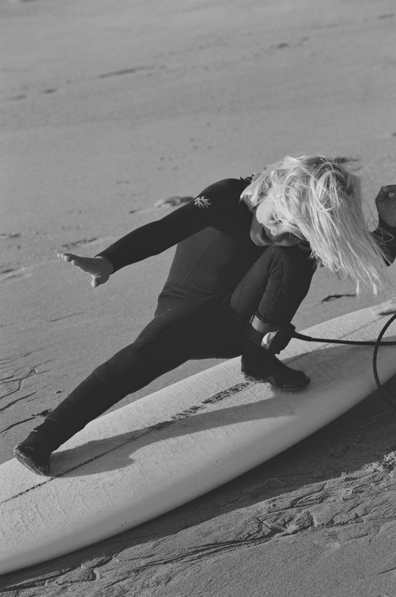Child in a Black wetsuit lying on a surfboard on a sandy beach