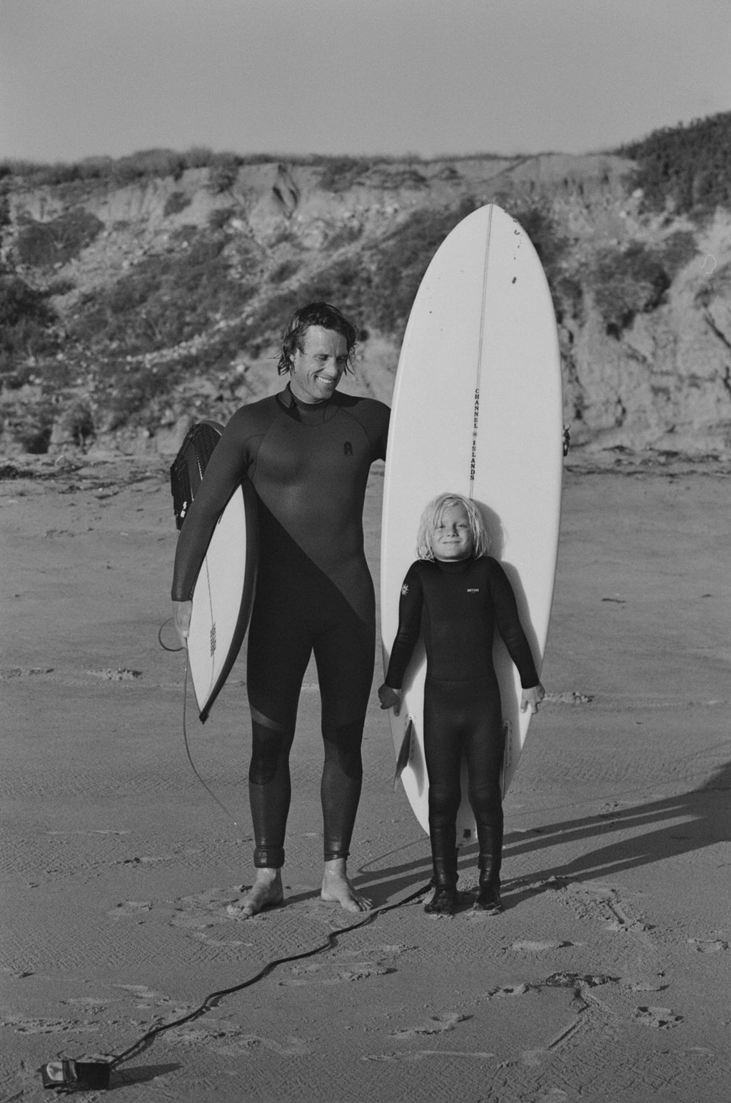 Two people in wetsuits holding surfboards on a beach with cliffs in the background.