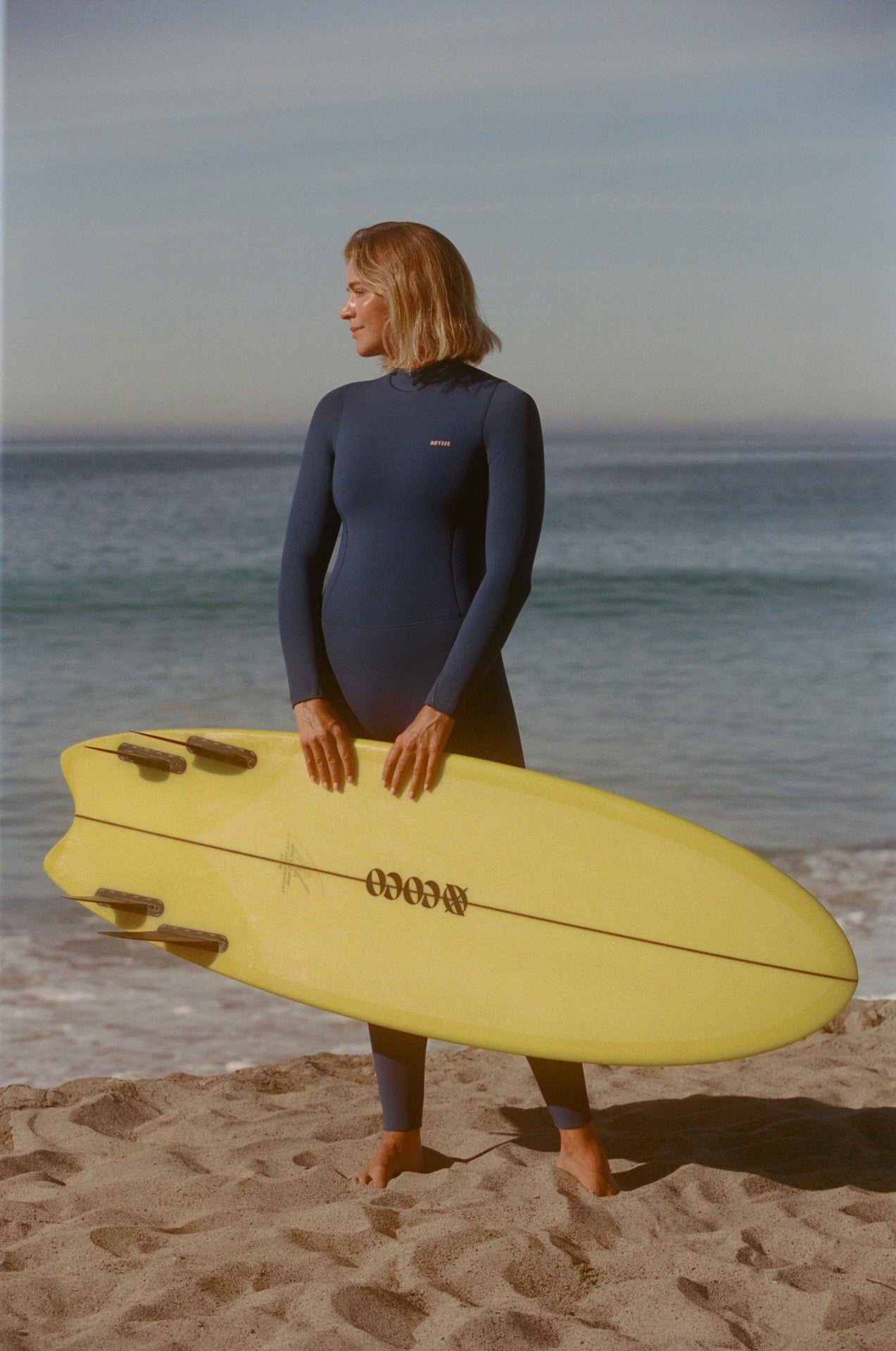 Woman in a wetsuit holding a yellow surfboard on a beach
