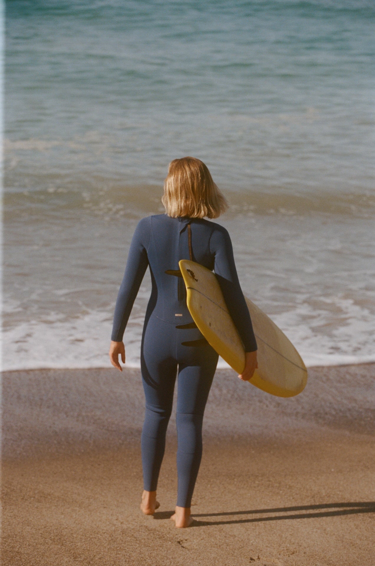 Person in a wetsuit walking on a beach with a surfboard under their arm.