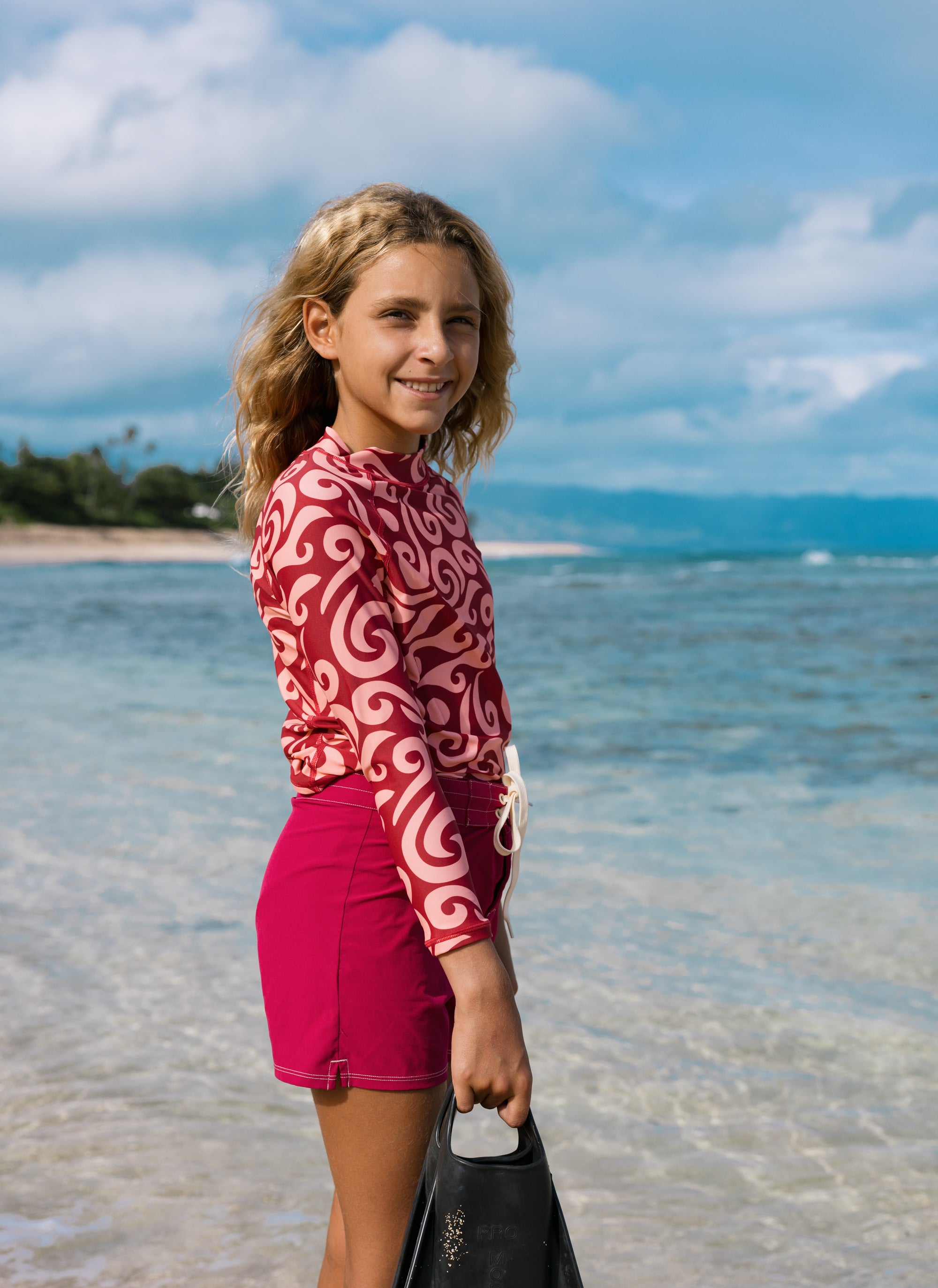A young girl stands on a beach shoreline wearing a red and white patterned long-sleeve swim shirt and matching pink swim shorts, holding a black flip-flop. The background shows blue ocean water, distant land, and a cloudy sky.