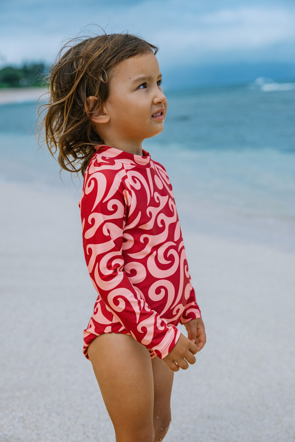 A young child wearing a red and white long-sleeve swimsuit with a bold swirl pattern, standing on a sandy beach with the ocean in the background.