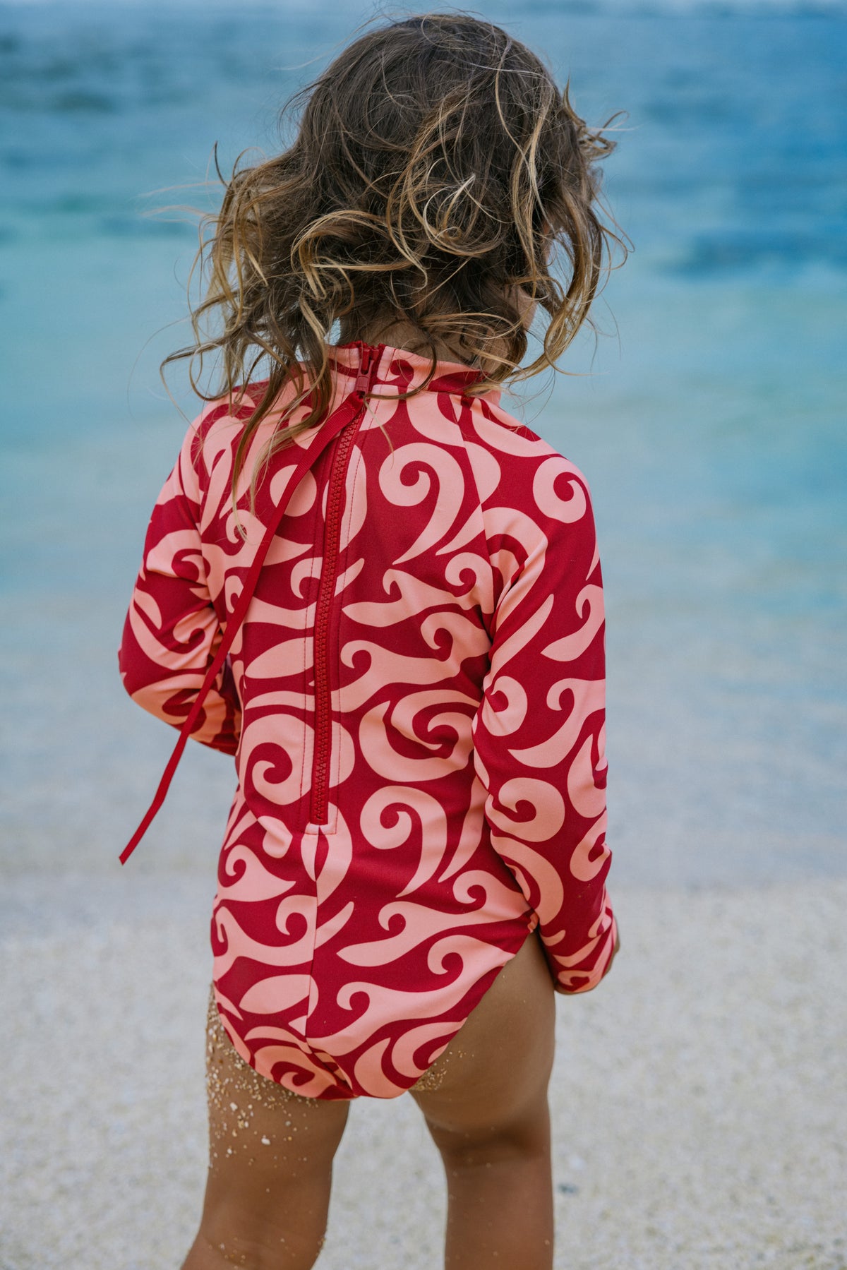 A young child viewed from behind on a beach, wearing a red long-sleeve swim cover-up with a pink tribal swirl pattern and a tie-neck design.