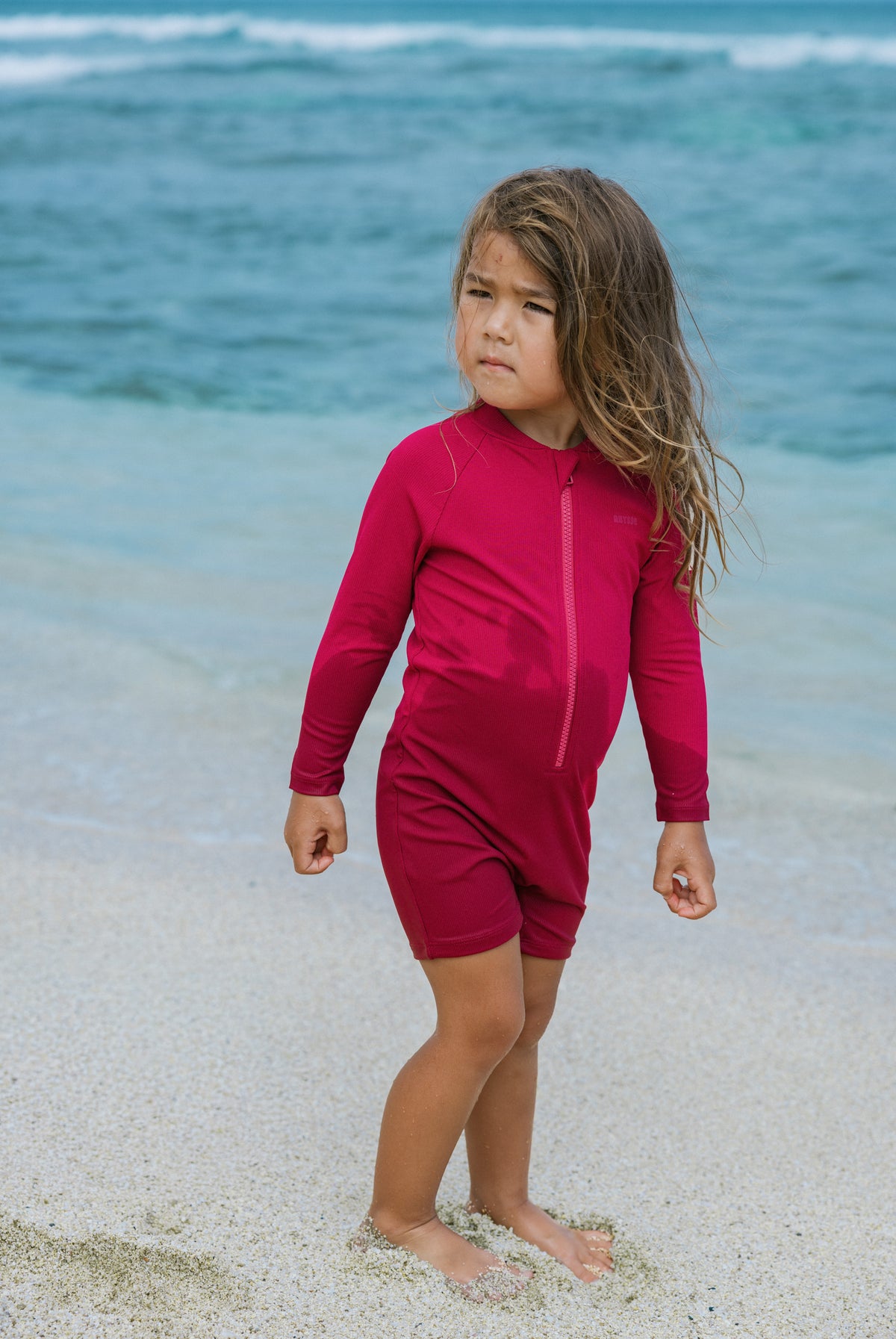 A young girl with long light brown hair stands on a sandy beach wearing a bright magenta-pink long-sleeve swim rash guard with a front zipper and matching swim shorts, with the ocean in the background.