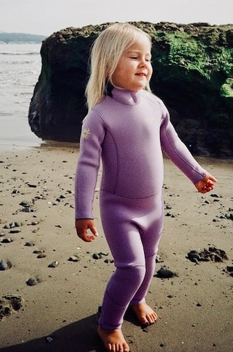Child in a purple wetsuit standing on a beach with rocks and water in the background