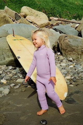 Child in a purple wetsuit standing on a rocky beach with a surfboard.