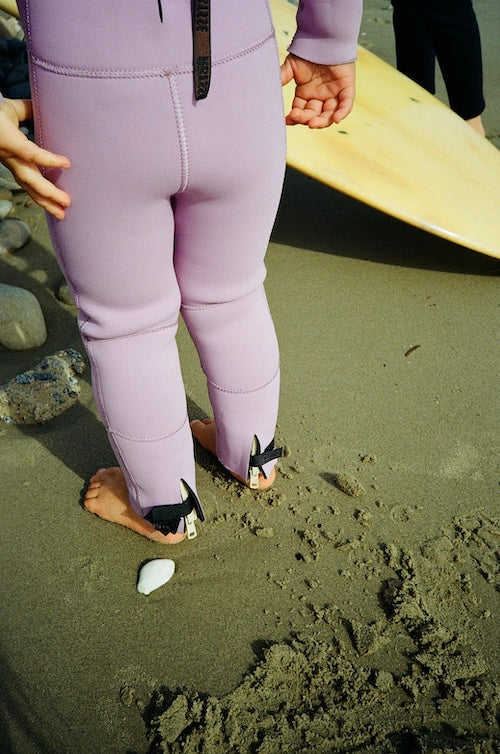Person wearing a purple wetsuit on a beach with a surfboard in the background.