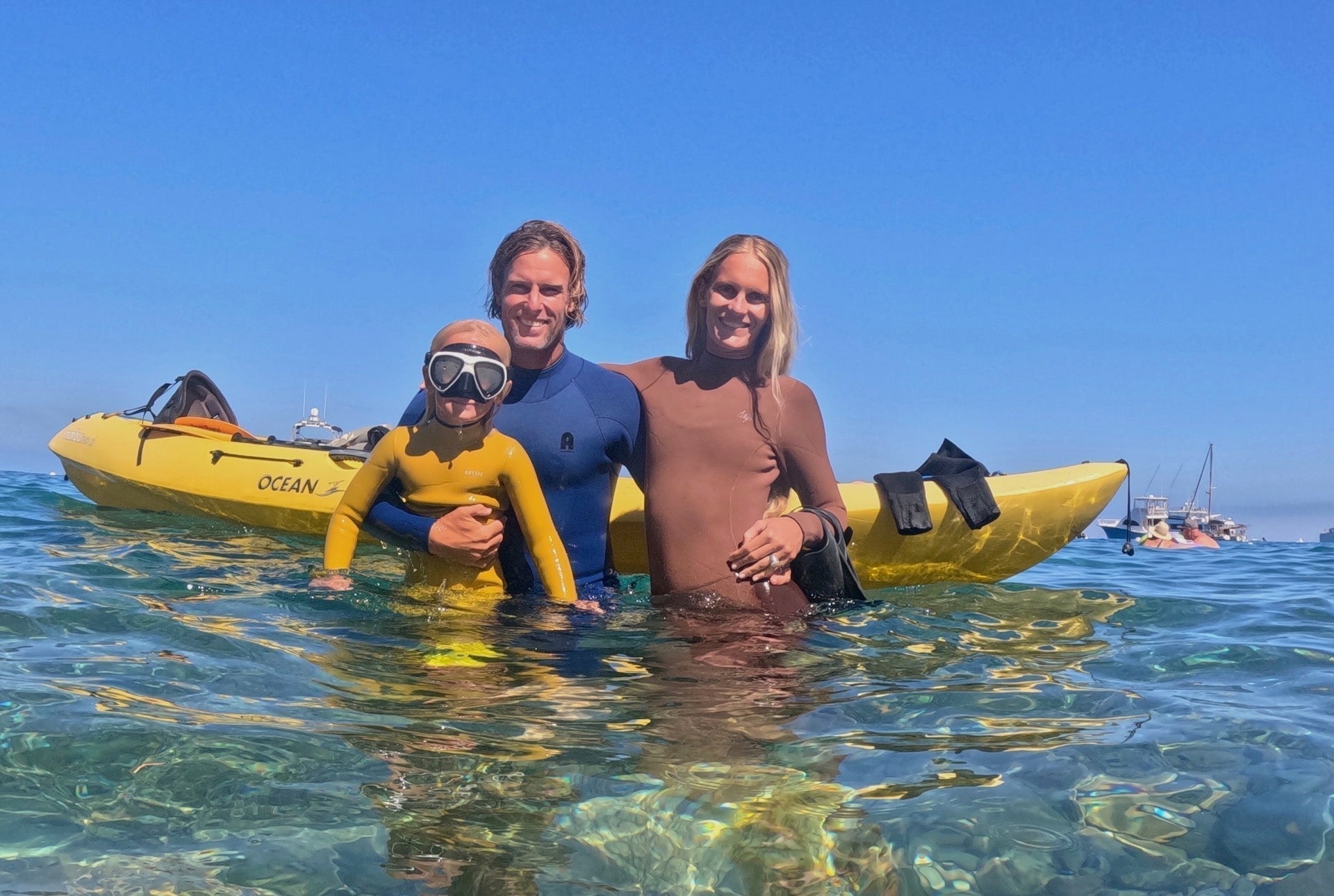 Family in Abysse wetsuits in the ocean — child wearing yellow Minis wetsuit with snorkel mask, adults in blue and reef brown wetsuits, kayak in background