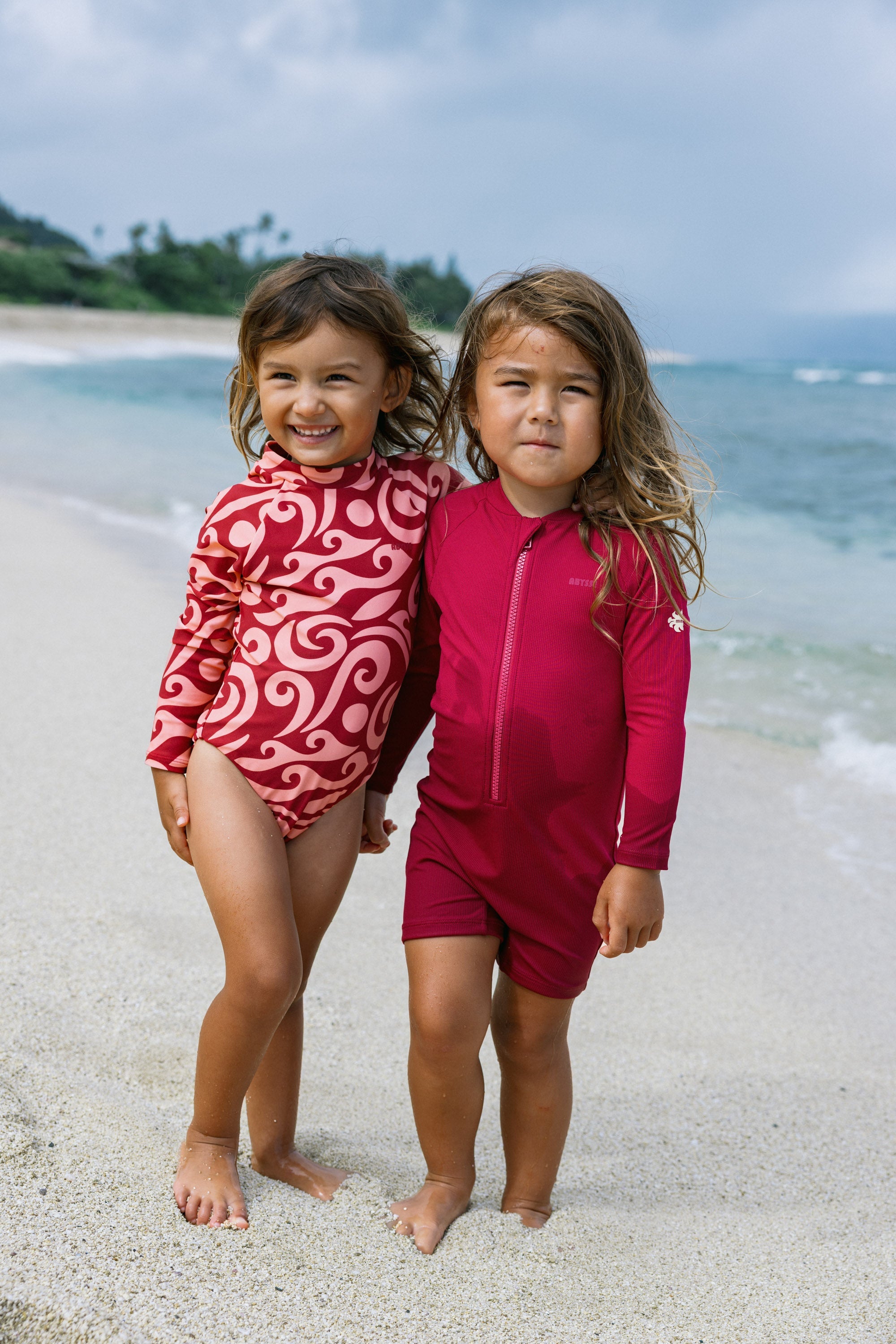 Two young girls standing on a sandy beach wearing matching long-sleeve swimwear sets. The girl on the left wears a red and white swirl patterned one-piece, and the girl on the right wears a solid dark pinkish-red one-piece with a front zipper.
