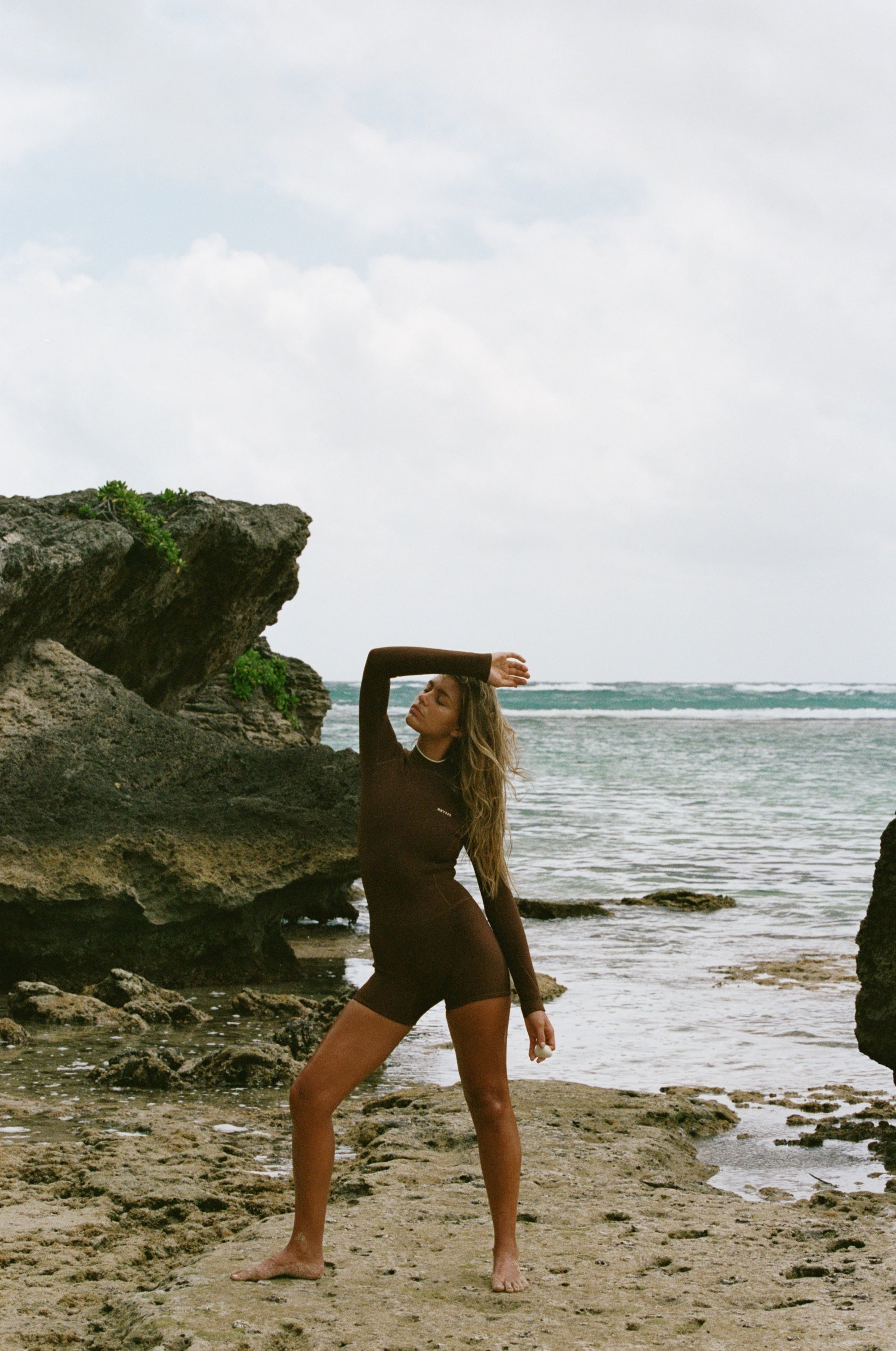 A woman in a dark brown full-body wetsuit poses on a rocky beach with the ocean and cloudy sky in the background.