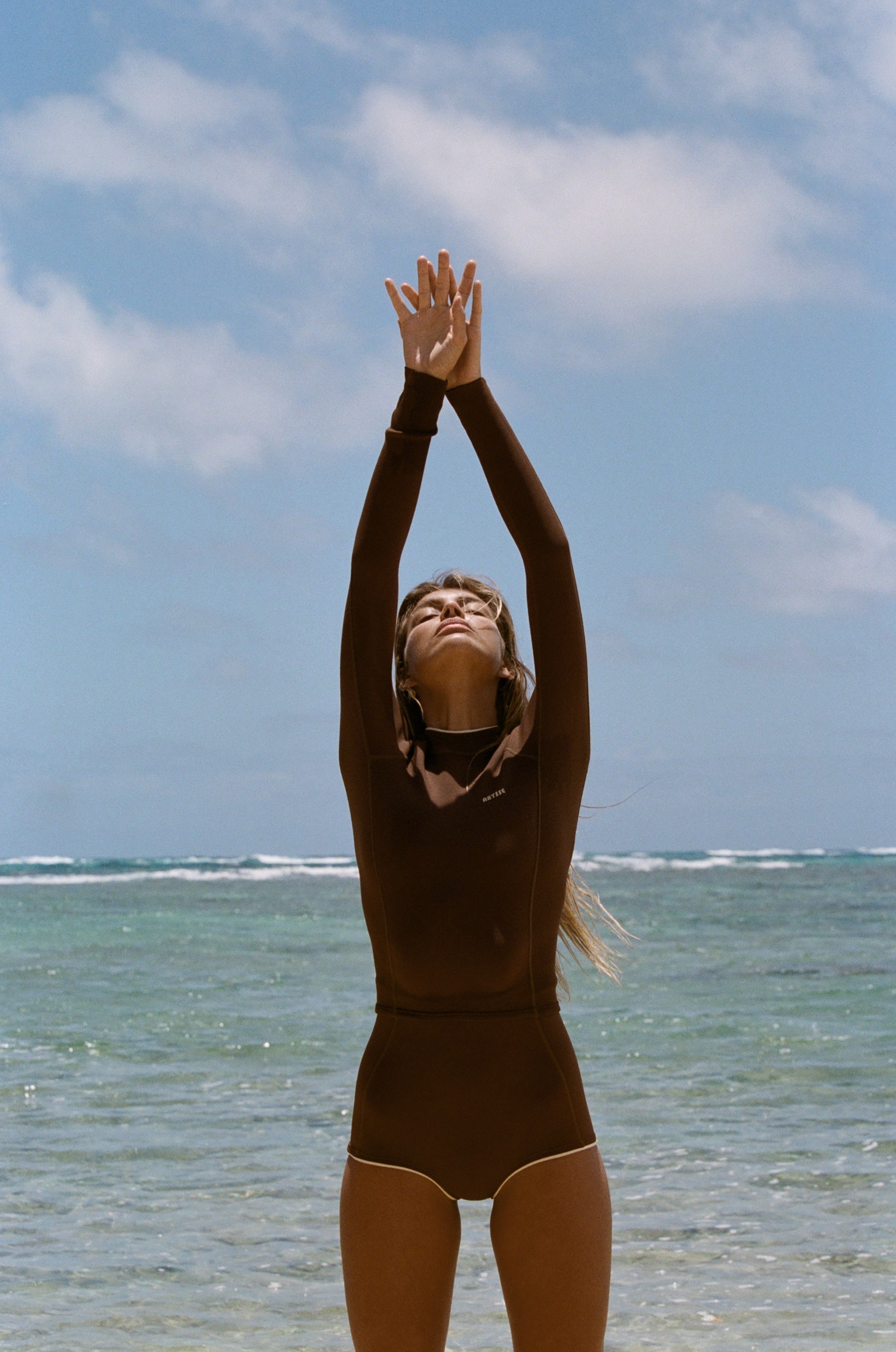 A woman in a dark brown full-body swimsuit or wetsuit stands on a beach with her arms raised above her head, looking upward.