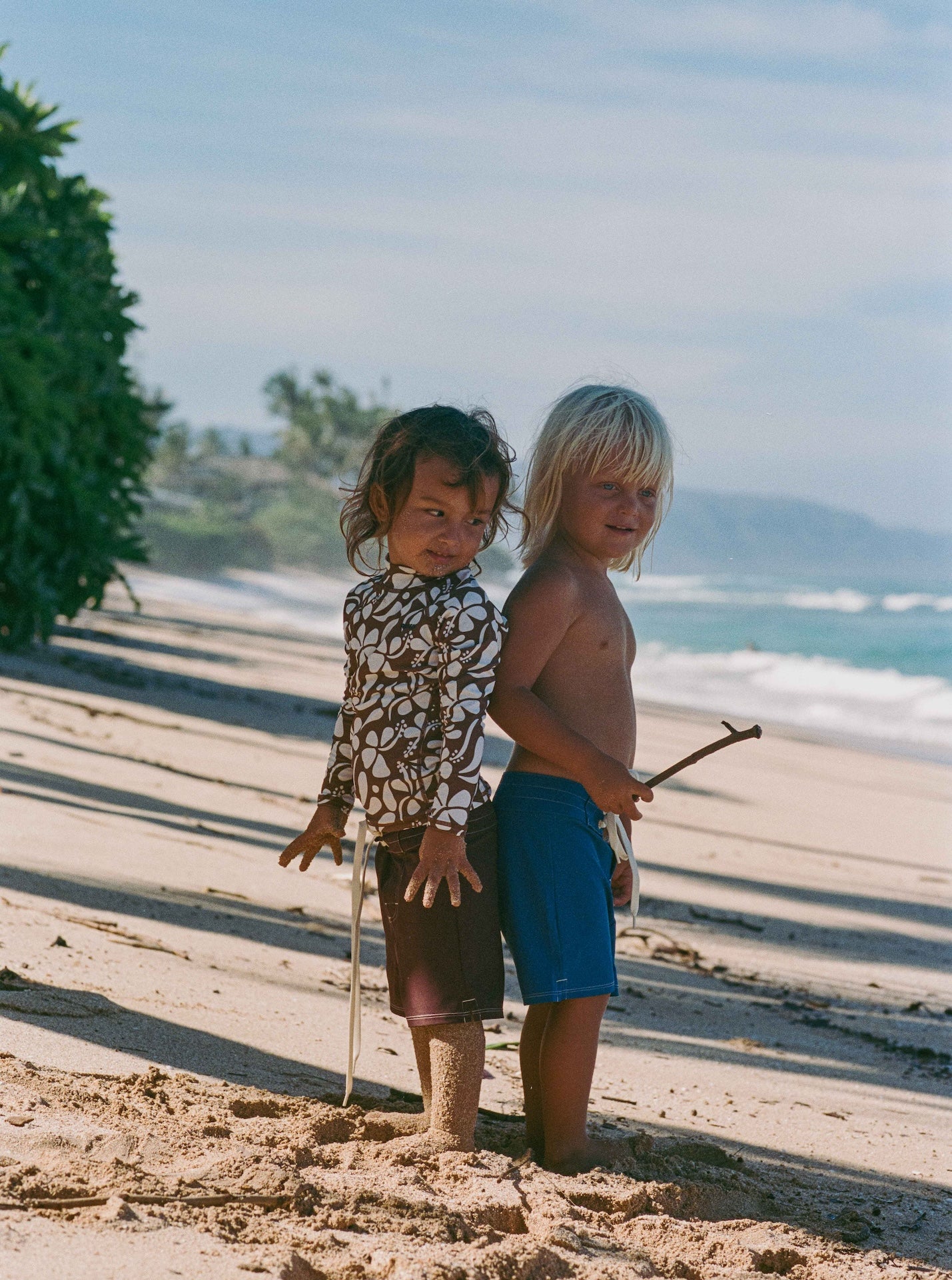 Two children standing on a sandy beach with ocean and sky in the background.