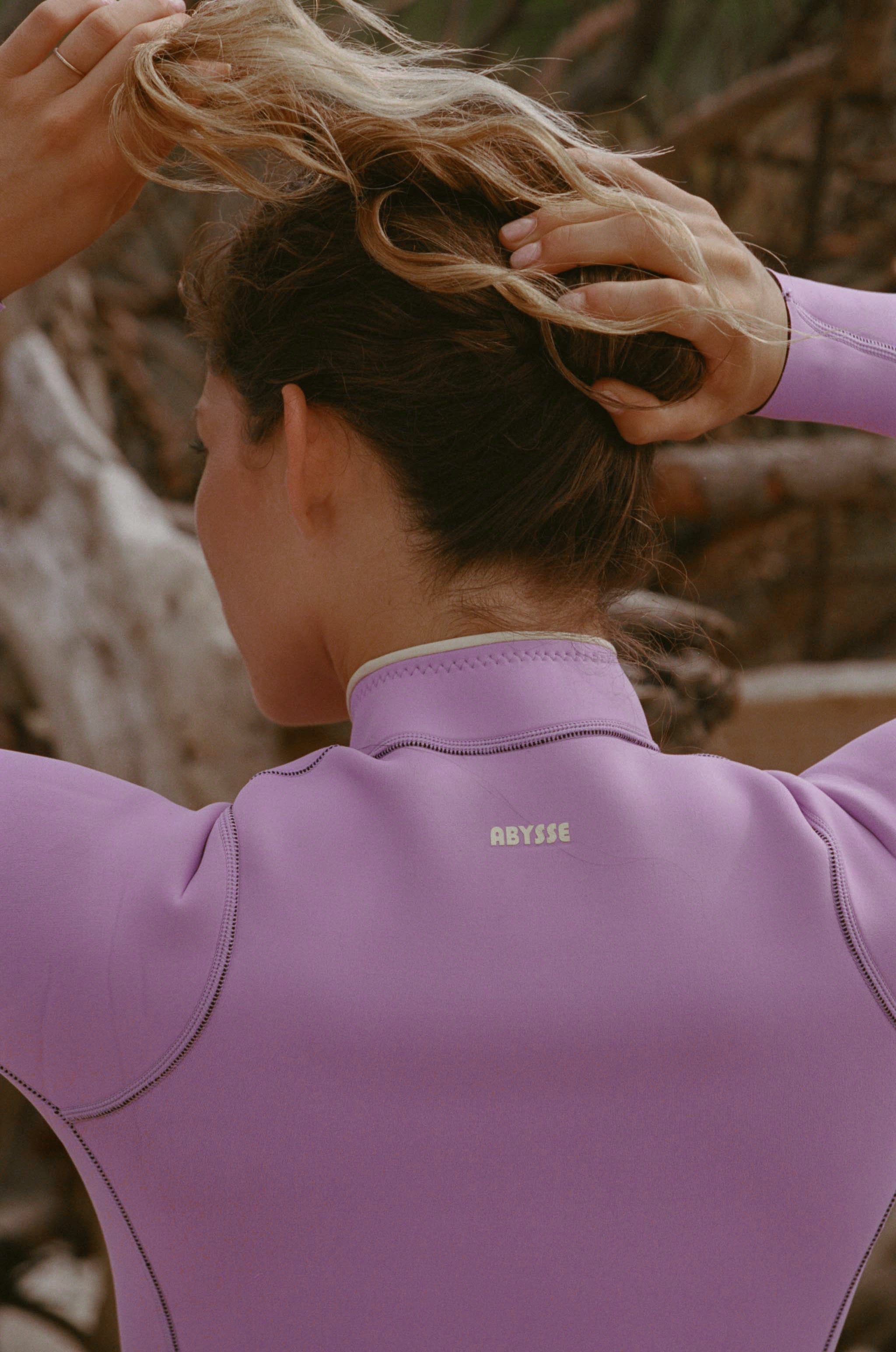 A woman wearing a lavender-purple wetsuit, seen from behind as she adjusts her hair.