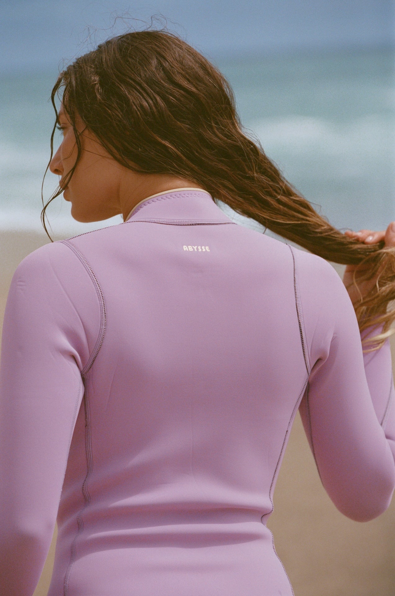 A woman on a beach wearing a light purple long-sleeve wetsuit, shown from behind with her hair in the air. The brand name 'NATIS' is printed in small white letters on the upper back.
