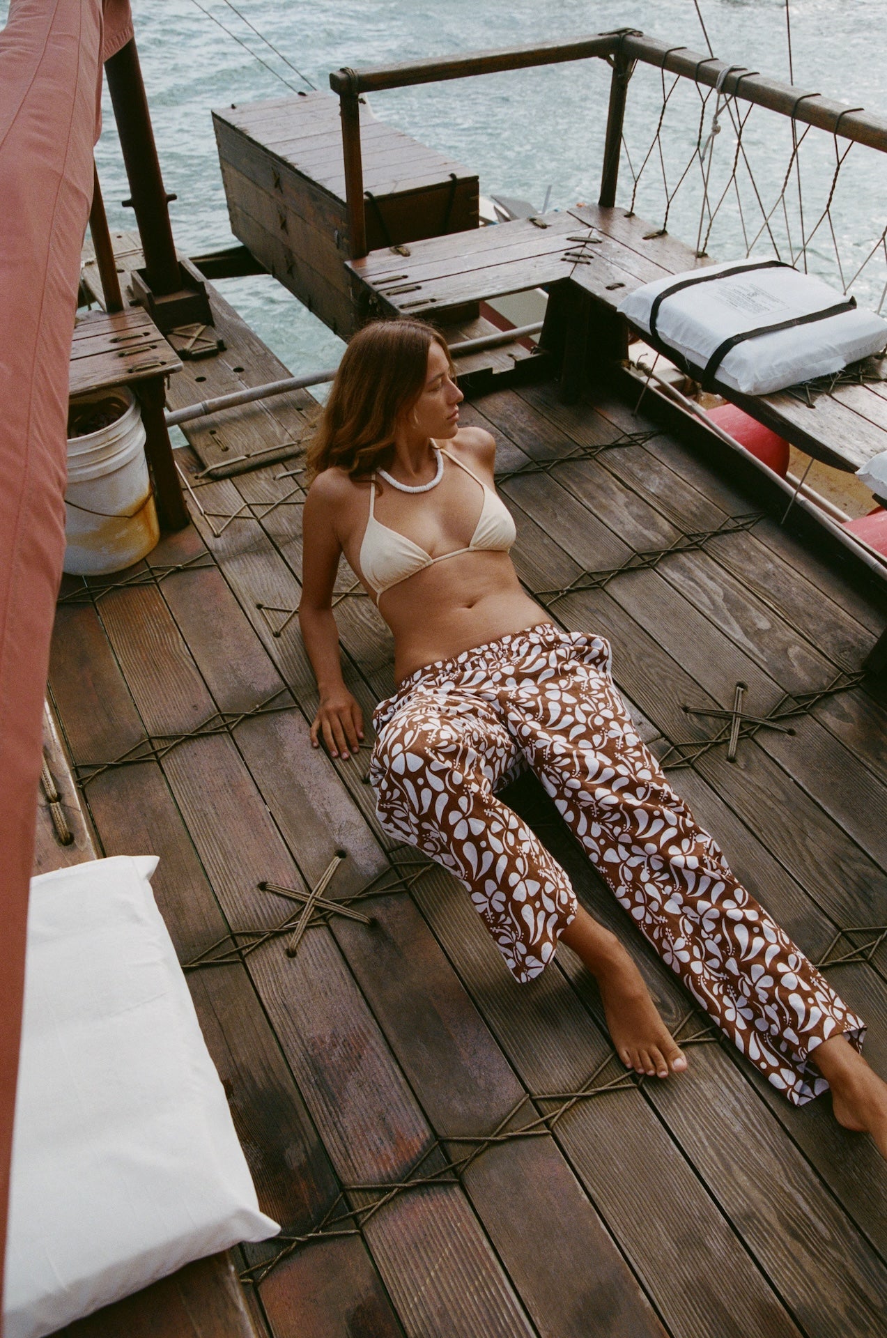 Woman in a bikini and patterned pants sitting on a wooden dock by the water.