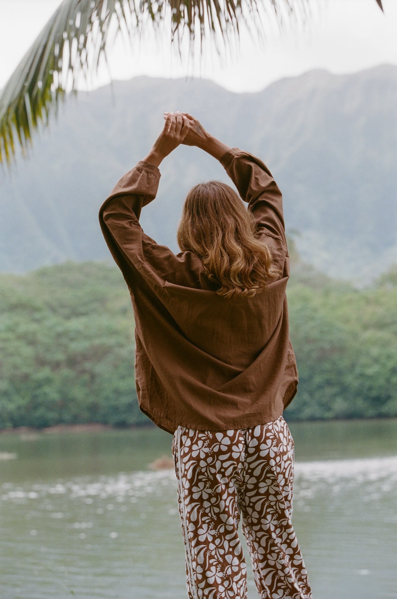 Person in a brown shirt and patterned pants standing by a lake with mountains in the background