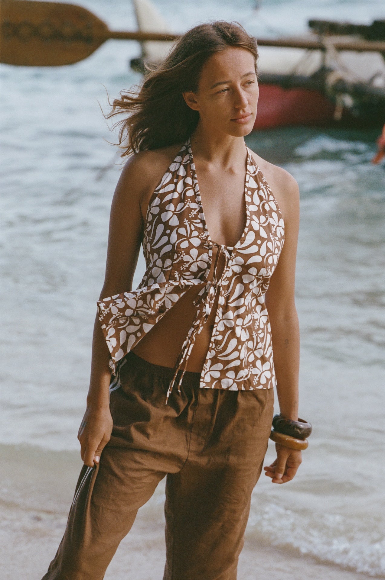 Woman in a patterned top and brown pants standing on a beach with water and a boat in the background.