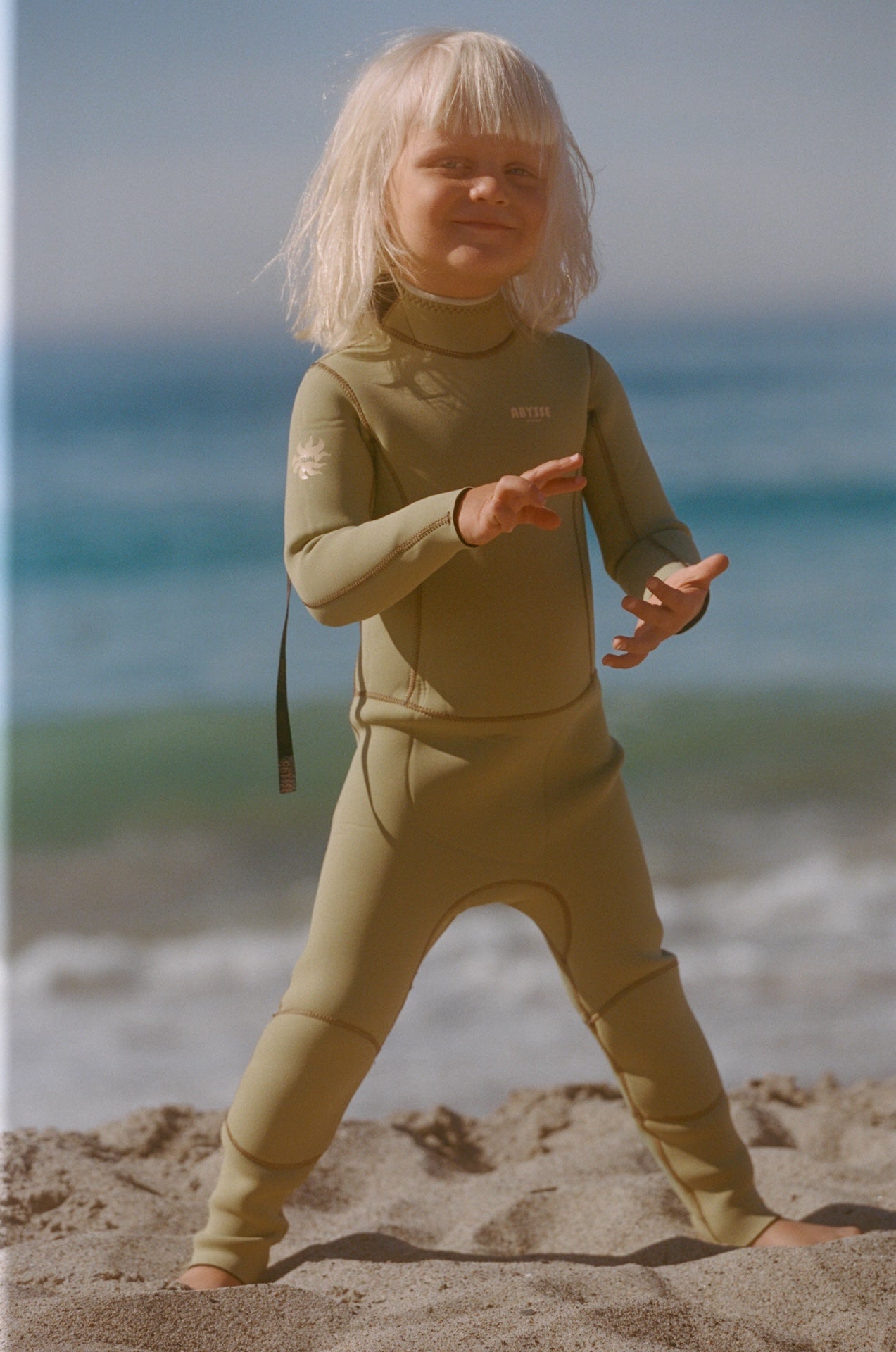 Child in a wetsuit standing on a beach with ocean in the background