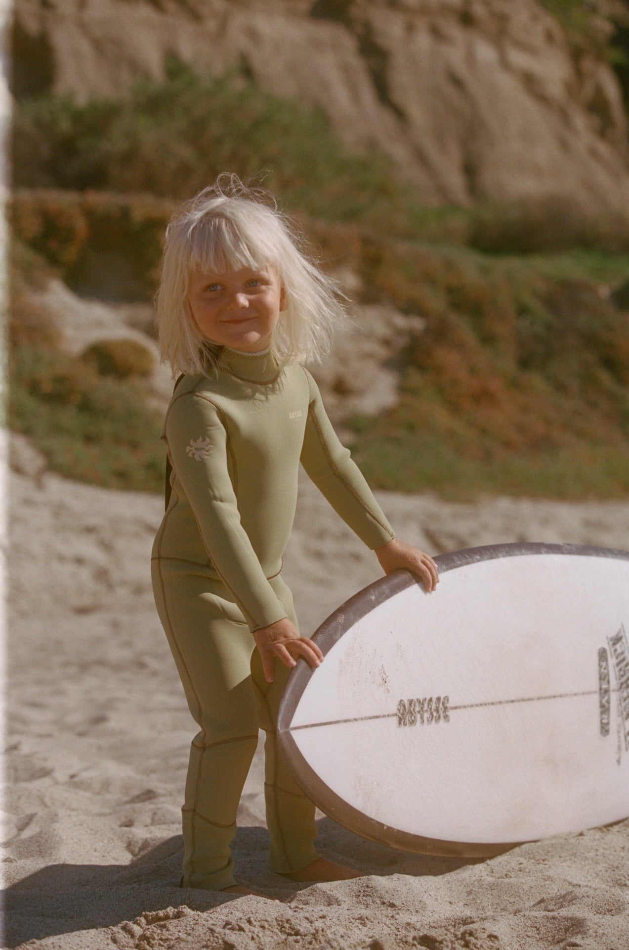 Child in a green wetsuit holding a white surfboard on a sandy beach with cliffs in the background.