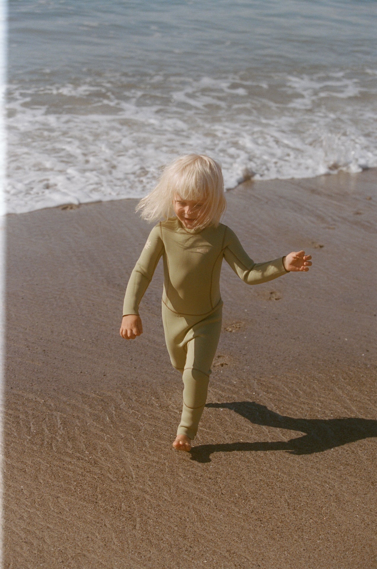 Child in a green outfit running on a sandy beach with ocean waves in the background
