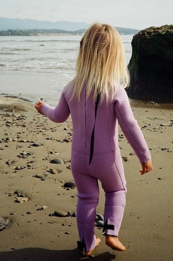 Person in a purple wetsuit walking on a beach with ocean and rocks in the background