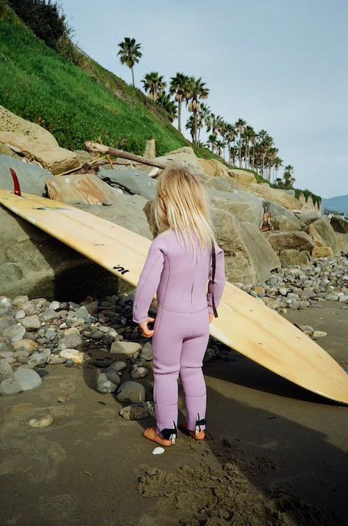 Person in a purple wetsuit holding a surfboard on a rocky beach with palm trees in the background